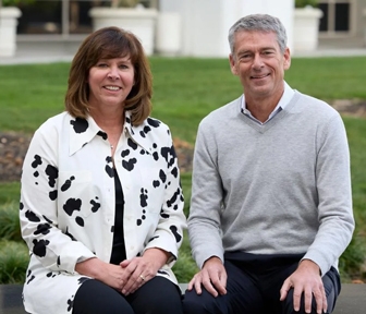 Joelle and Michael Hurlston seated on a concrete bench in front of a grassy area in front of a campus building.