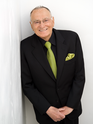 Jan Shrem smiling and leaning against a white wall with hands clasped wearing a black suit, green tie and green pocket square.